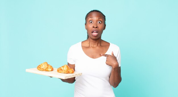 mirando conmocionado y sorprendido con la boca bien abierta, apuntando a sí mismo y sosteniendo una bandeja de croissants