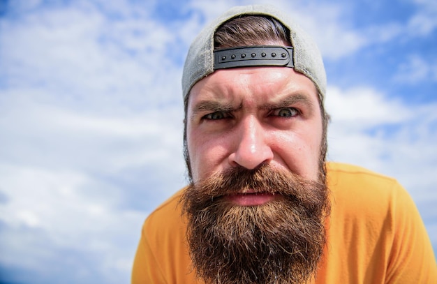 Mirando directamente delante de él Hombre brutal con barba larga y mirada seria Hombre barbudo con gorra hipster de moda al aire libre Hipster en ropa de moda callejera en cielo nublado Cool hipster