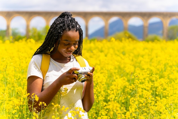 Mirando las fotos en una cámara vintage una chica étnica negra con trenzas un viajero en un campo de flores amarillas