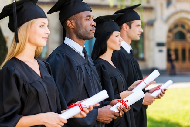 Mirando hacia un futuro brillante. cuatro graduados universitarios con sus diplomas y mirando hacia otro lado mientras están parados en una fila frente a la universidad