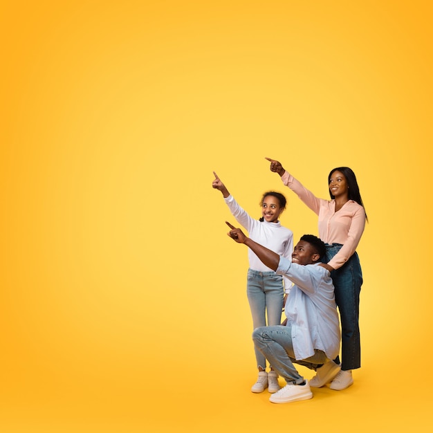 Mire allí Feliz familia afroamericana de tres personas apuntando al espacio libre posando sobre una foto de estudio de fondo amarillo