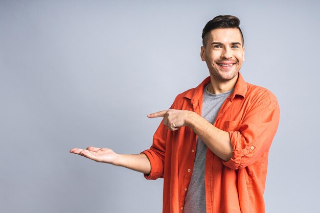 Mire allí Feliz joven apuesto en casual apuntando hacia el espacio de copias y sonriendo mientras está de pie aislado sobre fondo gris blanco