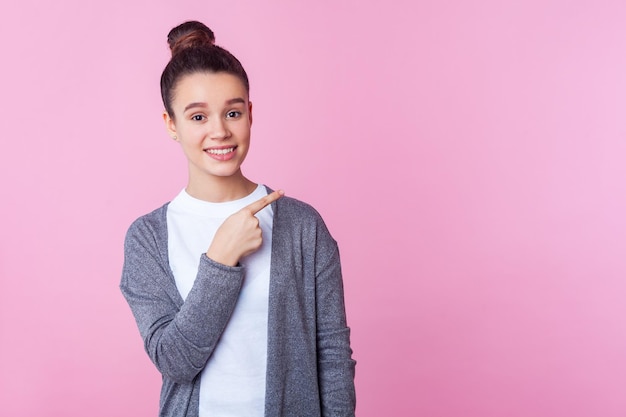 Mire el anuncio aquí Retrato de una encantadora adolescente feliz con peinado de moño en ropa casual apuntando a una pared vacía y una foto de estudio interior sonriente aislada en un espacio de copia de fondo rosa