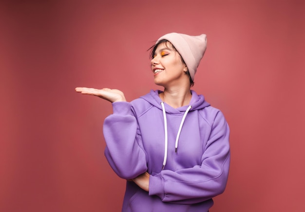 Mire aquí publicidad Retrato de una chica linda feliz en un suéter lila sosteniendo un espacio vacío en sus palmas una pared vacía para anunciar una foto de estudio aislada en un fondo rosa