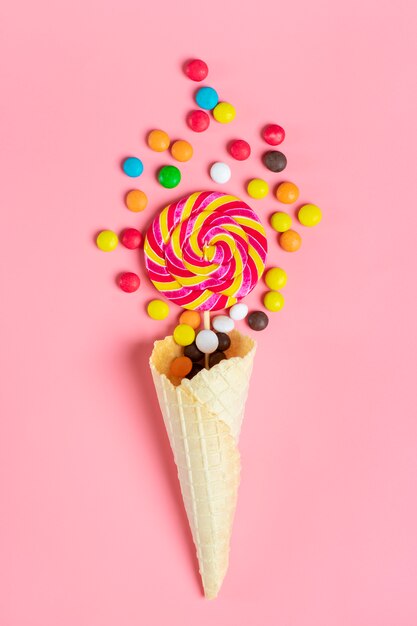 mischen Sie bunte Schokoladenbonbons, die aus Eiscremewaffelkegel auf rosa flacher Lage heraus verschüttet werden