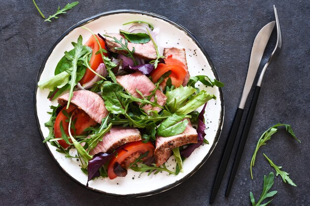 Mischen Sie Salat mit Tomaten und mittlerem Rindfleisch in einem Teller auf einem dunklen Hintergrund.