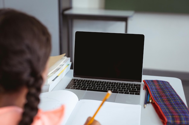 Mischlingsschülerin sitzt im Klassenzimmer und schreibt und benutzt Laptop, mit Platz für Kopien auf dem Bildschirm