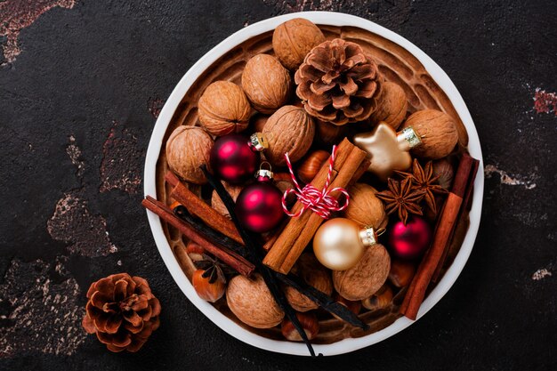 Mischung aus gesunden rohen Haselnüssen und Walnüssen, Zimtstangen, Anis, Vanille, Schokolade und Weihnachtsspielzeug in Keramikplatte auf brauner Betonoberfläche