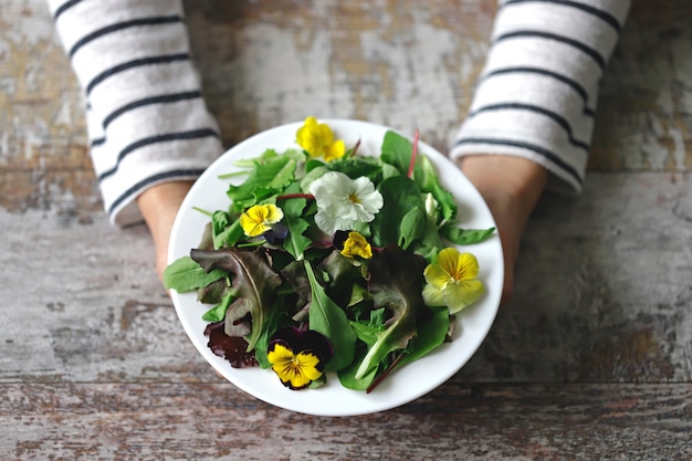 Mischung von Salaten mit Blumen auf einem weißen Teller, der von einem Mädchen gehalten wird