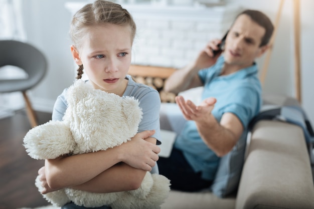 Miserável menina loira de olhos azuis segurando seu brinquedo e chorando enquanto seu pai falava ao telefone