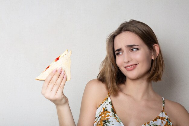 Misstrauische brünette Studentin, die ein Sandwich über einem grauen Hintergrund isst. Platz für Text