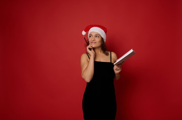 Misteriosa mujer bonita de raza mixta con sombrero de Santa sostiene el bloc de notas y mira pensativamente apuntando con lápiz en el espacio de la copia sobre fondo rojo. Conceptos de planificación de Navidad y año nuevo para publicidad.