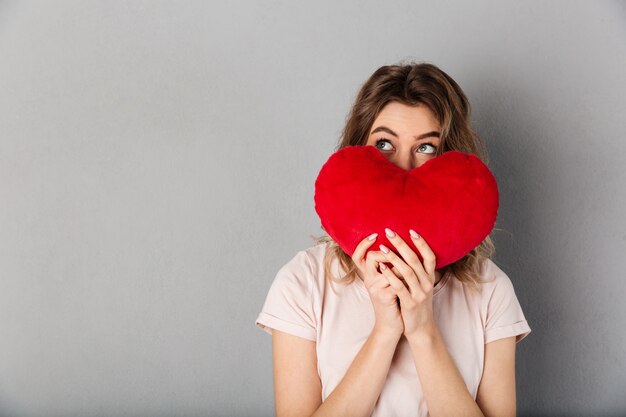 Foto misteriosa mujer en camiseta escondiéndose detrás de alegría corazón y mirando a otro lado sobre gris