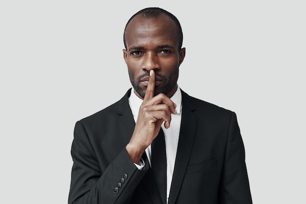 Misterioso joven africano en ropa formal manteniendo el dedo en los labios mientras está de pie contra la pared gris