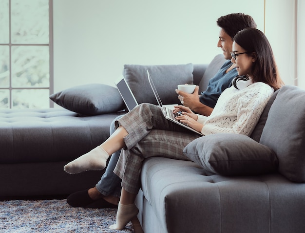 Misture os amantes da família de raça, marido caucasiano e mulher asiática, sentados na sala de estar trabalhando no sofá, usando o laptop, em gestos fáceis de relaxamento. conceito de trabalhar em casa e novo estilo de vida moderno normal.