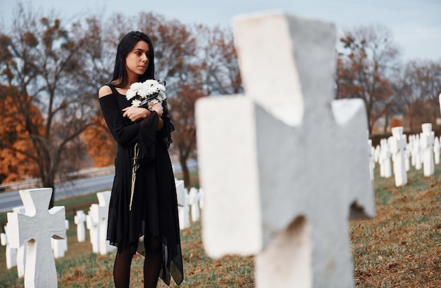 Mit Blumen in den Händen Junge Frau in schwarzer Kleidung, die den Friedhof mit vielen weißen Kreuzen besucht Konzeption von Beerdigung und Tod
