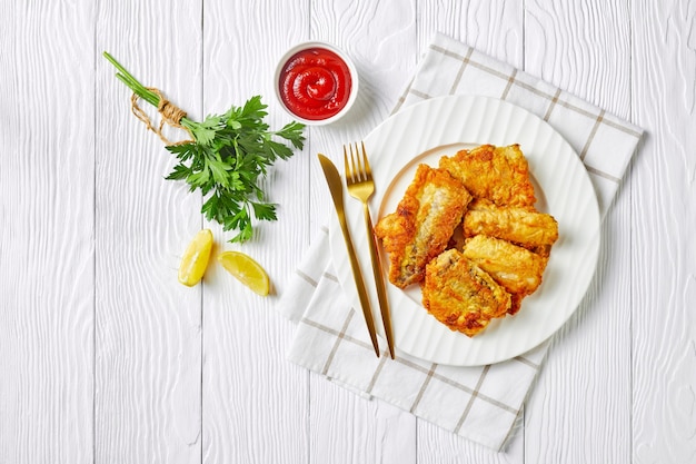 Mit Butter gebratenes Kabeljaufilet nach britischer Art, serviert auf einem weißen Teller mit Besteck, Tomatenketchup, frischer Petersilie auf einer weißen Holzoberfläche, Draufsicht, flache Lage