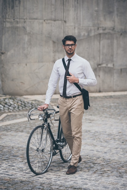 Mit dem Fahrrad zur Arbeit. Selbstbewusster junger Mann mit Brille, der wegschaut und die Hand auf seinem Fahrrad hält, während er im Freien geht