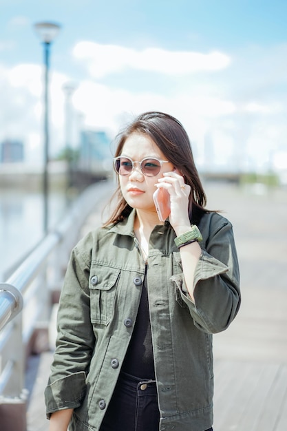 Mit dem Smartphone der jungen schönen asiatischen Frau mit Jacke und schwarzer Jeans, die im Freien posiert