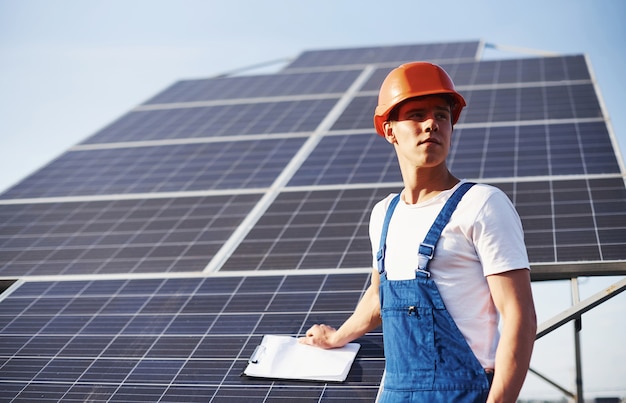 Mit Dokumenten. Männlicher Arbeiter in blauer Uniform im Freien mit Solarbatterien am sonnigen Tag.