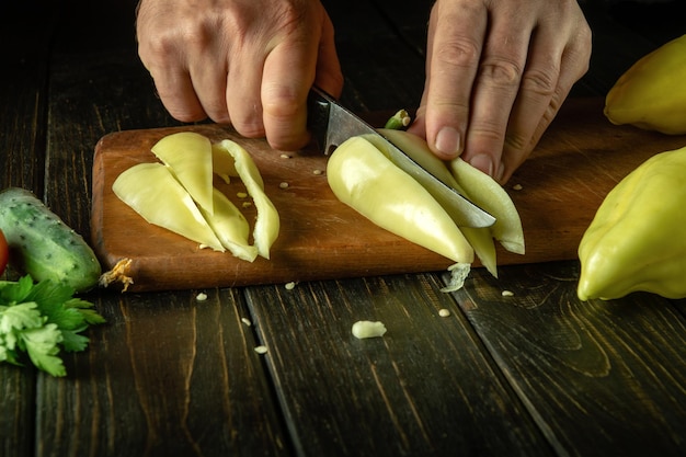 Mit einem Messer in den Händen eines Kochs Paprika für ein Gemüsegericht schneiden. Das Konzept der Gemüsediät. Dunkler Raum für Rezept oder Menü
