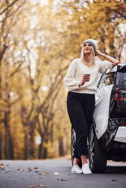 Mit einer Tasse Getränk in den Händen. Mädchen haben eine Herbstreise mit dem Auto. Modernes brandneues Auto im Wald.