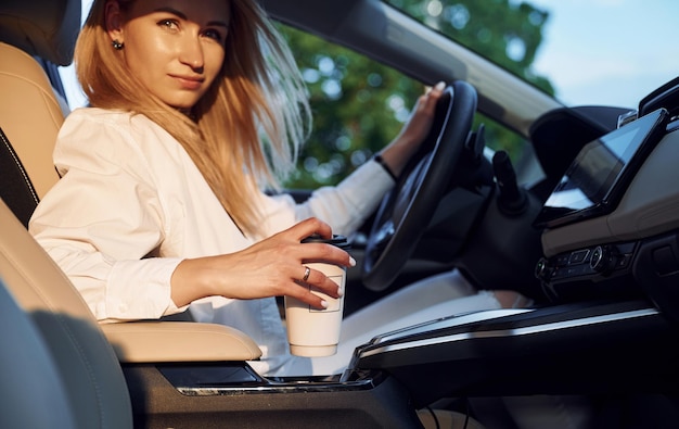 Mit einer Tasse Getränk ist eine junge Frau in weißer Kleidung tagsüber mit ihrem Elektroauto unterwegs