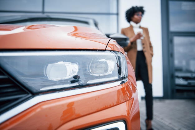 Mit einer Tasse Getränk. Junge Afroamerikanerin mit Brille steht draußen in der Nähe eines modernen Autos.