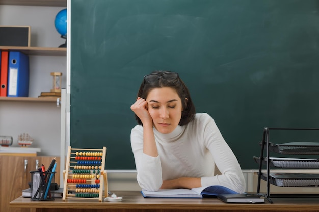 mit geschlossenen augen junge lehrerin sitzt am schreibtisch mit schulwerkzeugen im klassenzimmer