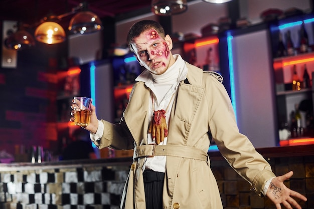 Mit getränk in der hand. porträt des mannes, der auf der thematischen halloween-party in zombie-make-up und -kostüm ist.