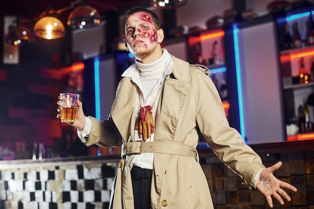 Mit Getränk in der Hand. Porträt des Mannes, der auf der thematischen Halloween-Party in Zombie-Make-up und -kostüm ist.