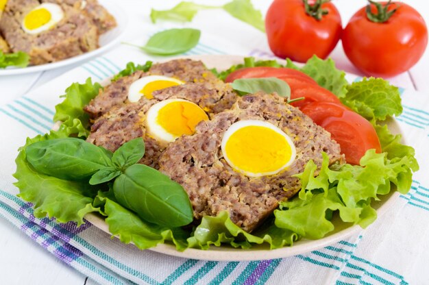 Mit Hackbraten gefülltes gekochtes Ei mit frischen Tomaten- und Salatblättern auf einem Teller auf einem weißen Holztisch.