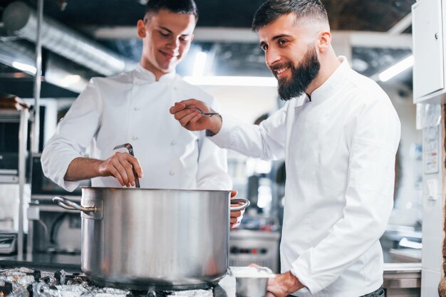 Mit Hilfe eines anderen Mitarbeiters Koch in weißer Uniform kocht Essen in der Küche Beschäftigter Tag bei der Arbeit