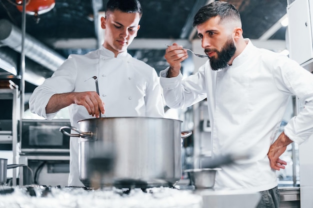 Mit Hilfe eines anderen Mitarbeiters Koch in weißer Uniform kocht Essen in der Küche Beschäftigter Tag bei der Arbeit