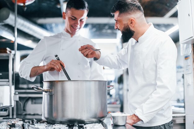 Mit Hilfe eines anderen Mitarbeiters Küchenchef in weißer Uniform, der in der Küche Essen kocht Beschäftigter Tag bei der Arbeit