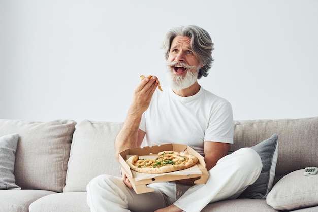 Mit leckerer Pizza Uhren TV-Show Älterer stilvoller moderner Mann mit grauem Haar und Bart drinnen