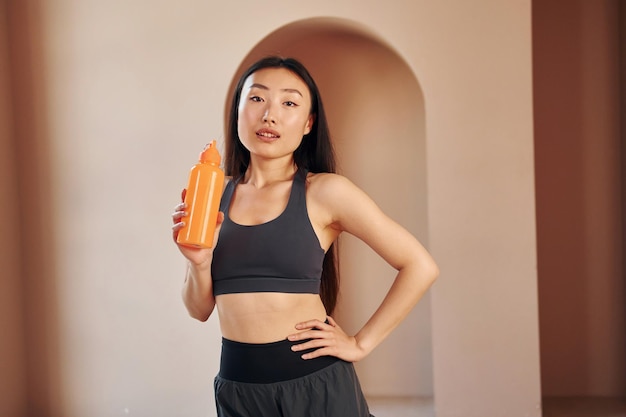 Mit orangefarbener Flasche Junge ernsthafte asiatische Frau, die drinnen steht