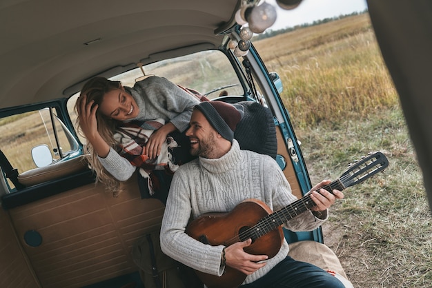 Mit seinem Herzen spielen. Hübscher junger Mann, der Gitarre für seine schöne Freundin spielt, während er im Minivan im Retro-Stil sitzt