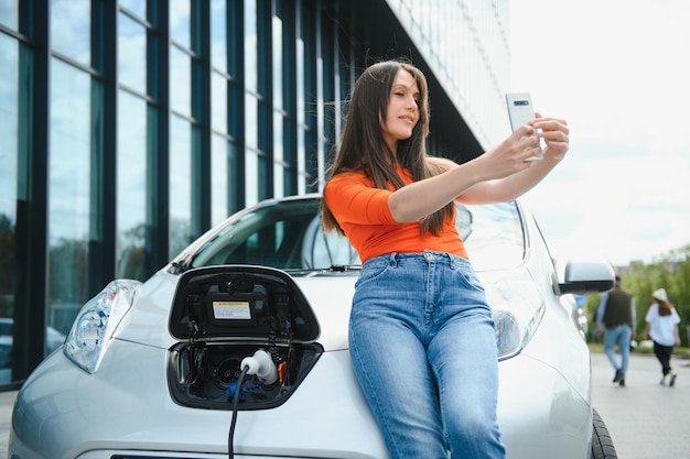 Mit Smartphone während des Wartens Frau an der Ladestation für Elektroautos tagsüber Brandneues Fahrzeug