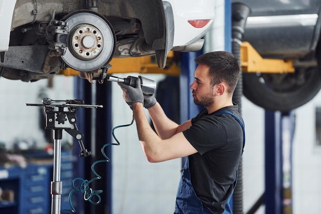 Mit Spezialwerkzeug Mann in Arbeitsuniform repariert weißes Auto im Innenbereich Konzeption des Autoservices