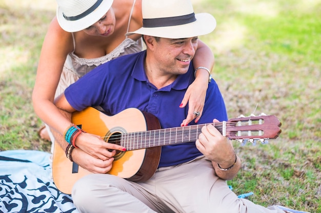 Mitad hombre adulto tocando la guitarra mientras la esposa abrazando desde atrás sentado en el jardín. Pareja amorosa pasar tiempo libre durante las vacaciones. Feliz pareja pasar tiempo libre tocando la guitarra al aire libre