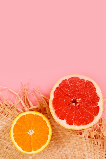 La mitad de pomelo y naranja y un sombrero de paja sobre un fondo rosa.