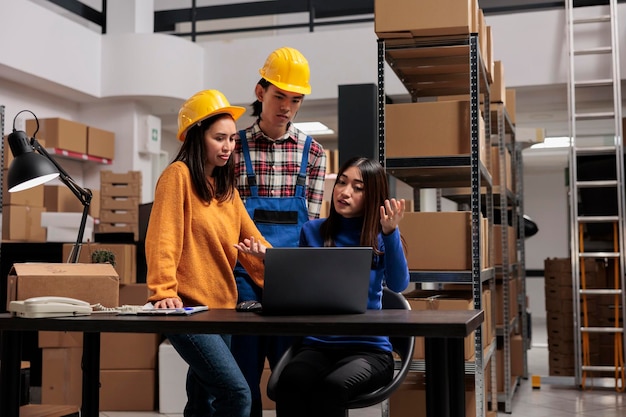 Mitarbeiter des asiatischen Vertriebszentrums überprüfen den Warenlieferplan auf dem Laptop. Lagerleiterin sitzt am Schreibtisch und erklärt Kollegen im industriellen Lagerraum die Logistikkette