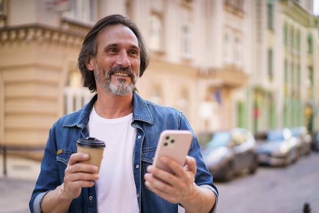 Mithilfe der Navigation steht ein reifer Mann mit seinem Mobiltelefon auf der Straße einer europäischen Stadt. Reisekonzept. Hübscher Mann mittleren Alters genießt seinen Coffee to go und reist in lässiger Kleidung durch die Altstadt