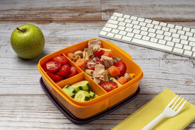 Mittagessen am Arbeitsplatz gesunde Pasta mit Thunfisch, Kirschtomaten, Karotten, Gurke in der Brotdose auf dem Arbeitstisch. Hauptnahrungsmittel für Bürokonzept