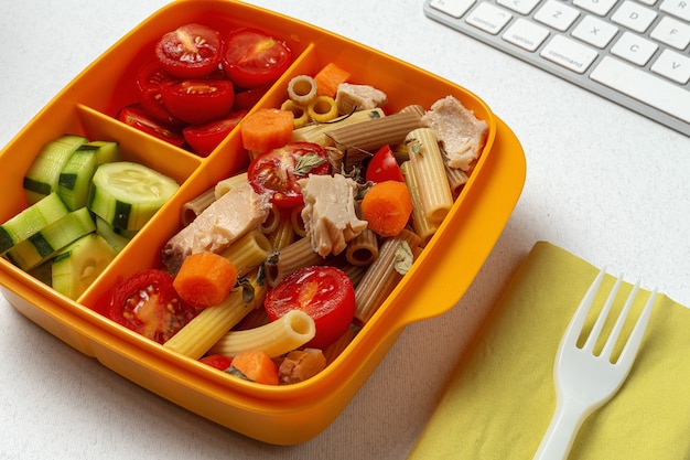 Mittagessen am Arbeitsplatz gesunde Pasta mit Thunfisch, Kirschtomaten, Karotten, Gurke in der Brotdose auf dem Arbeitstisch. Hauptnahrungsmittel für Bürokonzept