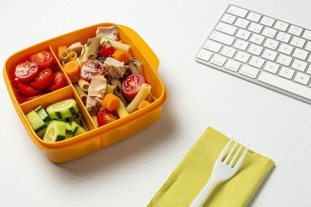 Mittagessen am Arbeitsplatz gesunde Pasta mit Thunfisch, Kirschtomaten, Karotten, Gurke in der Brotdose auf dem Arbeitstisch. Hauptnahrungsmittel für Bürokonzept