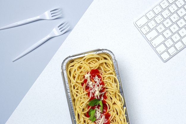 Mittagessen am Arbeitsplatz hausgemachte und gesunde Spaghetti mit Tomaten und Käse auf Arbeitstisch mit Computertastatur. Hauptnahrungsmittel für Bürokonzept. Essen zum Mitnehmen