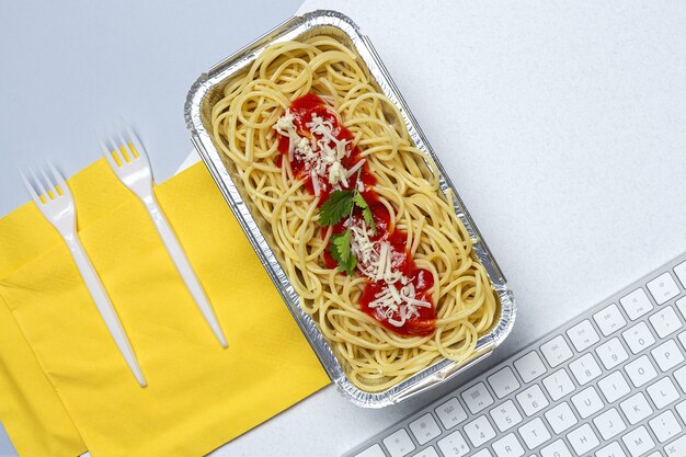 Mittagessen am Arbeitsplatz hausgemachte und gesunde Spaghetti mit Tomaten und Käse auf Arbeitstisch mit Computertastatur. Hauptnahrungsmittel für Bürokonzept. Essen zum Mitnehmen