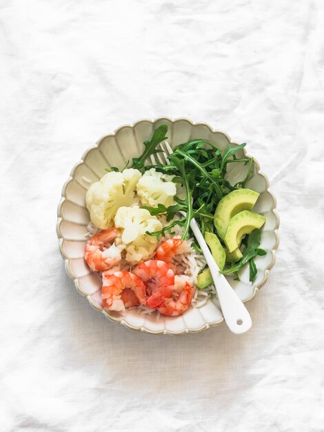 Mittagessen mit Reis, Garnelen, gekochtem Blumenkohl, Arugula, Avocado auf einem Teller auf einem hellen Hintergrund